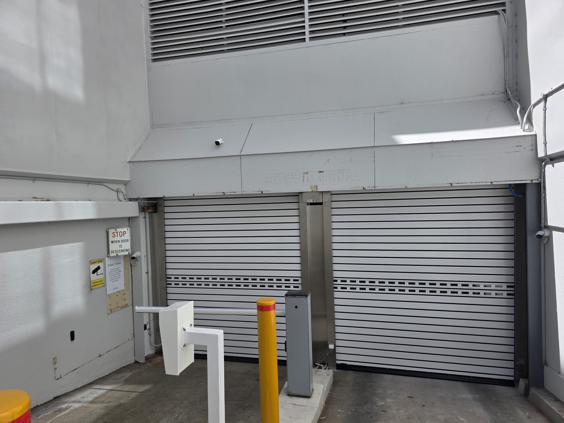 Paired grey high-speed car-park doors viewed from the vehicle ramp, with a barrier, controller and safety bollard