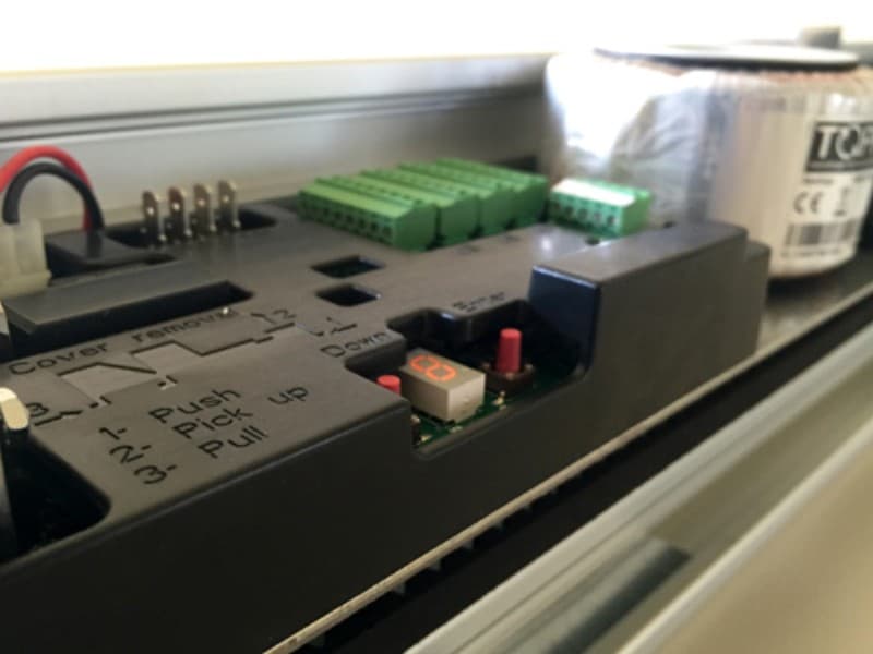 Close-up view of an automatic sliding door control unit, featuring wiring terminals, green input connectors, and labelled function buttons for push, pick up, and pull operations.