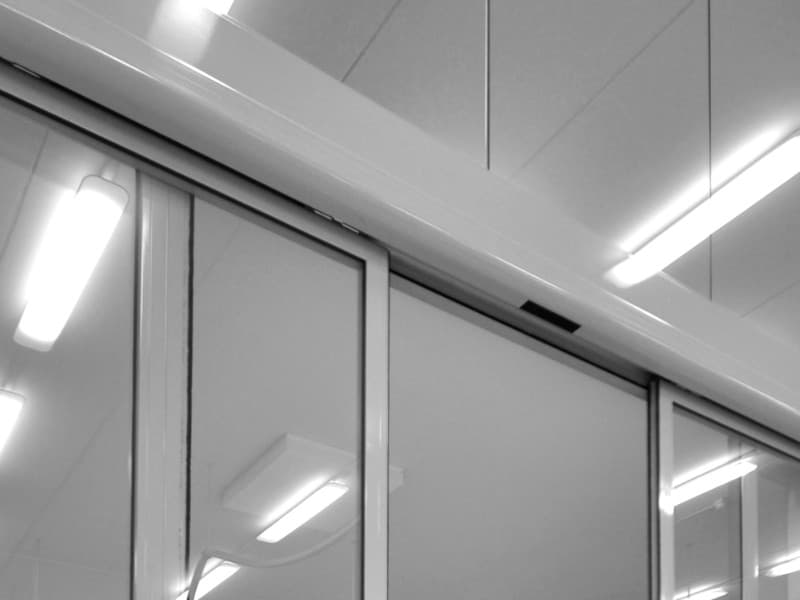Frameless automatic sliding glass doors installed in a commercial interior, with overhead motion sensor and integrated aluminium track system beneath a ceiling with fluorescent lighting.