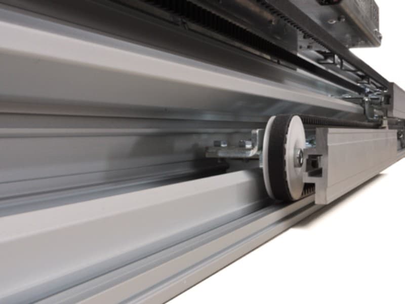 Close-up view of an automatic sliding door track system, highlighting the roller mechanism and toothed belt used for smooth and controlled door movement.