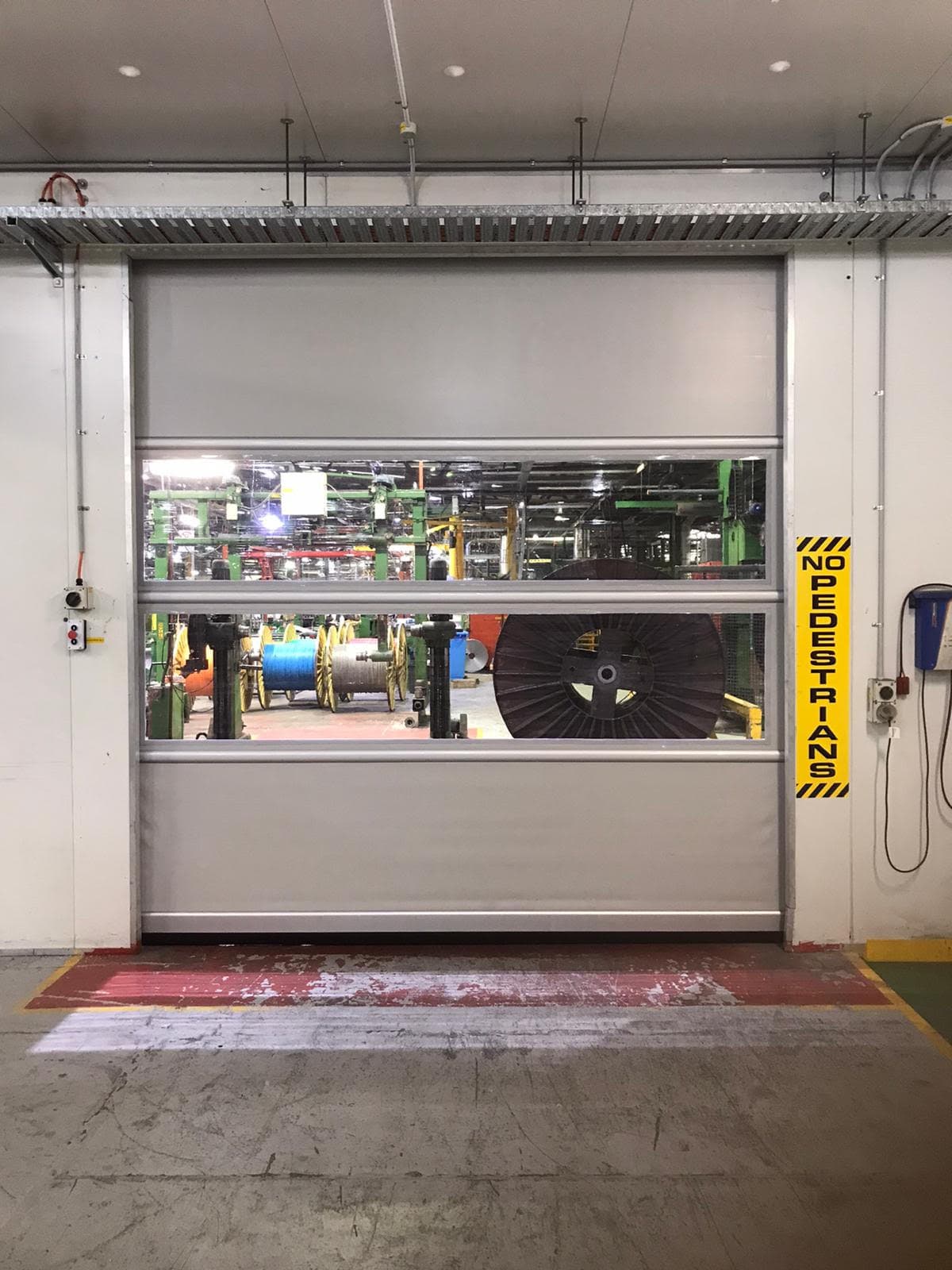 High-speed door at cables depot with transparent vision panels showing industrial cable spools and manufacturing equipment. A yellow 'NO PEDESTRIANS' safety sign is mounted on the right side, and the doorway has red and yellow floor markings.