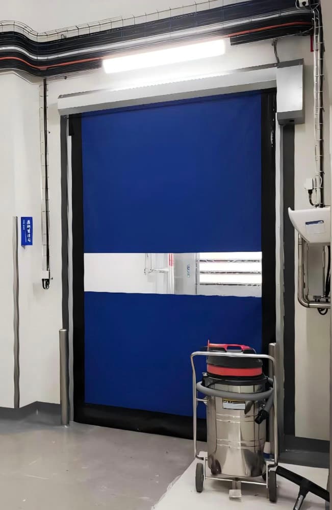 Blue high-speed door with a transparent vision panel installed in a clean room environment, with a stainless steel cleaning cart in the foreground.