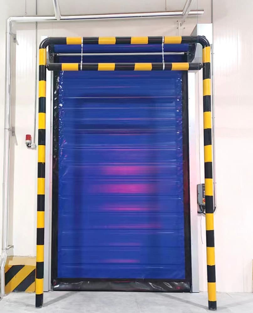 Photo of a blue high-speed roller door with black and yellow striped safety bollards mounted on both sides, installed in a white industrial doorway with a yellow hazard marking at the base.