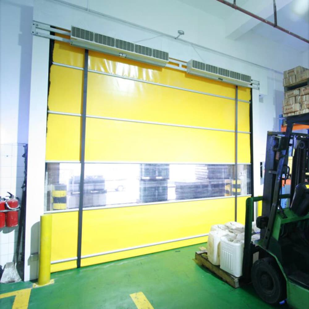 Photo of a yellow high-speed roller door with a transparent vision panel installed in a warehouse with green epoxy flooring, yellow floor markings, and a forklift visible to the right. A fire extinguisher and storage containers are also visible in the space.