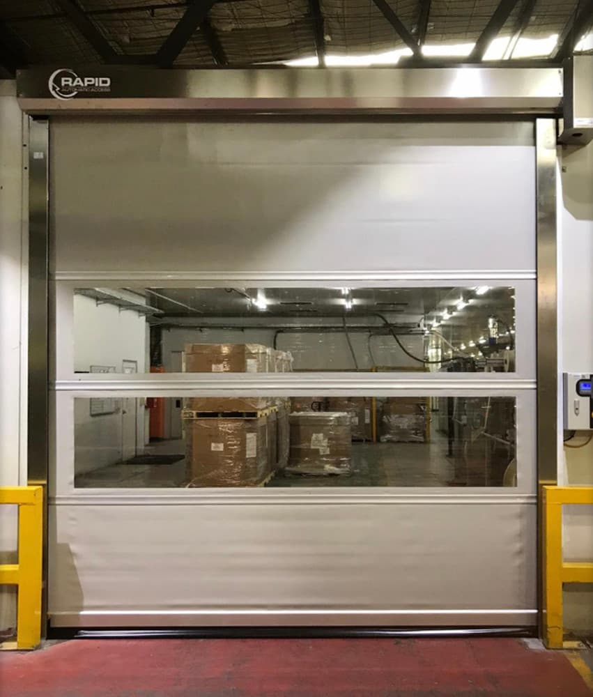 Photo of a high-speed door with two transparent vision panels showing stacked cardboard boxes in a warehouse interior. The door frame has yellow safety barriers on both sides, and the Rapid logo is visible on the top header. A red floor marking is visible at the base.