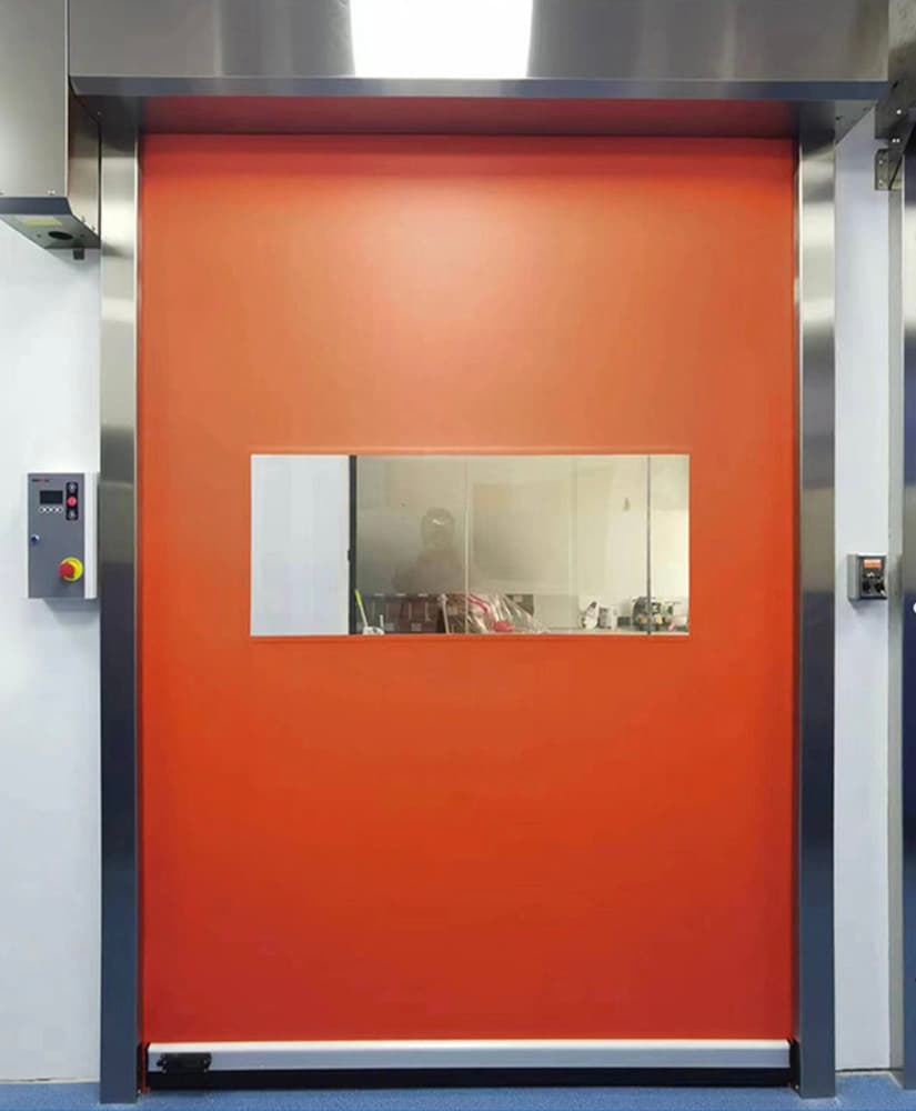 Photo of a red high-speed door with a horizontal vision panel in a stainless steel frame, installed in a clean room environment. Control panels are mounted on both sides, and the door features a blue floor base and white wall surroundings.