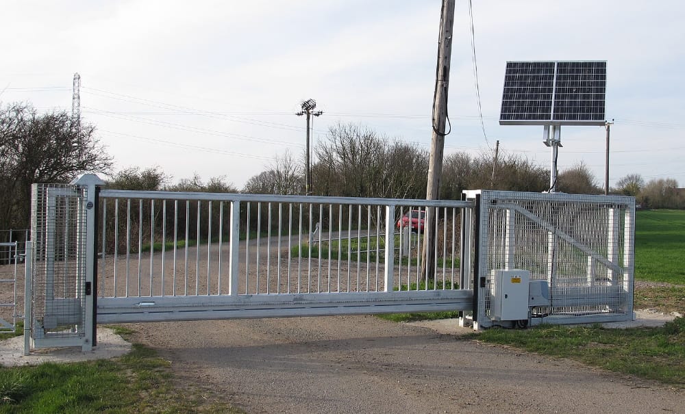 Solar powered sliding gate opener with solar panels installed beside a rural driveway gate.