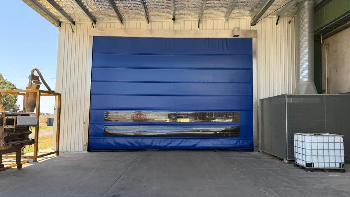 Blue high-speed door installed beneath a covered external loading bay with storage tanks and a concrete apron