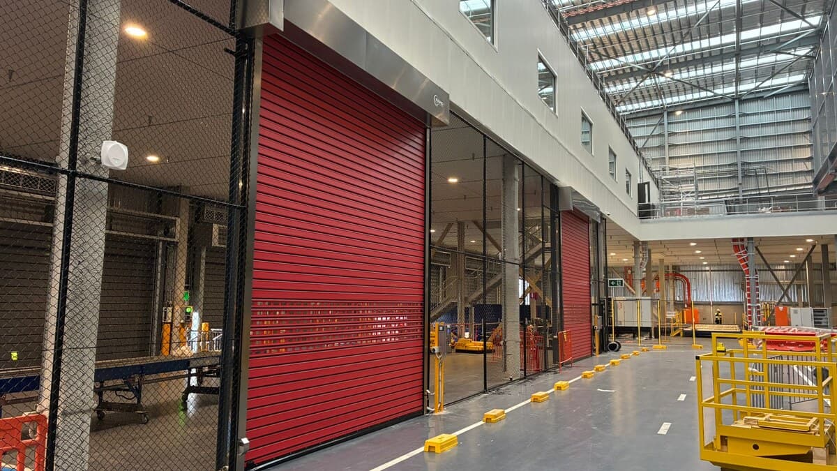 Red high-speed door installed beside a marked traffic lane and mesh storage enclosures inside a large warehouse