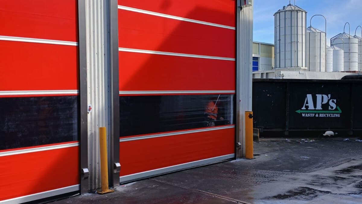 Two red high-speed doors installed at a waste and recycling facility with wet pavement and silos visible