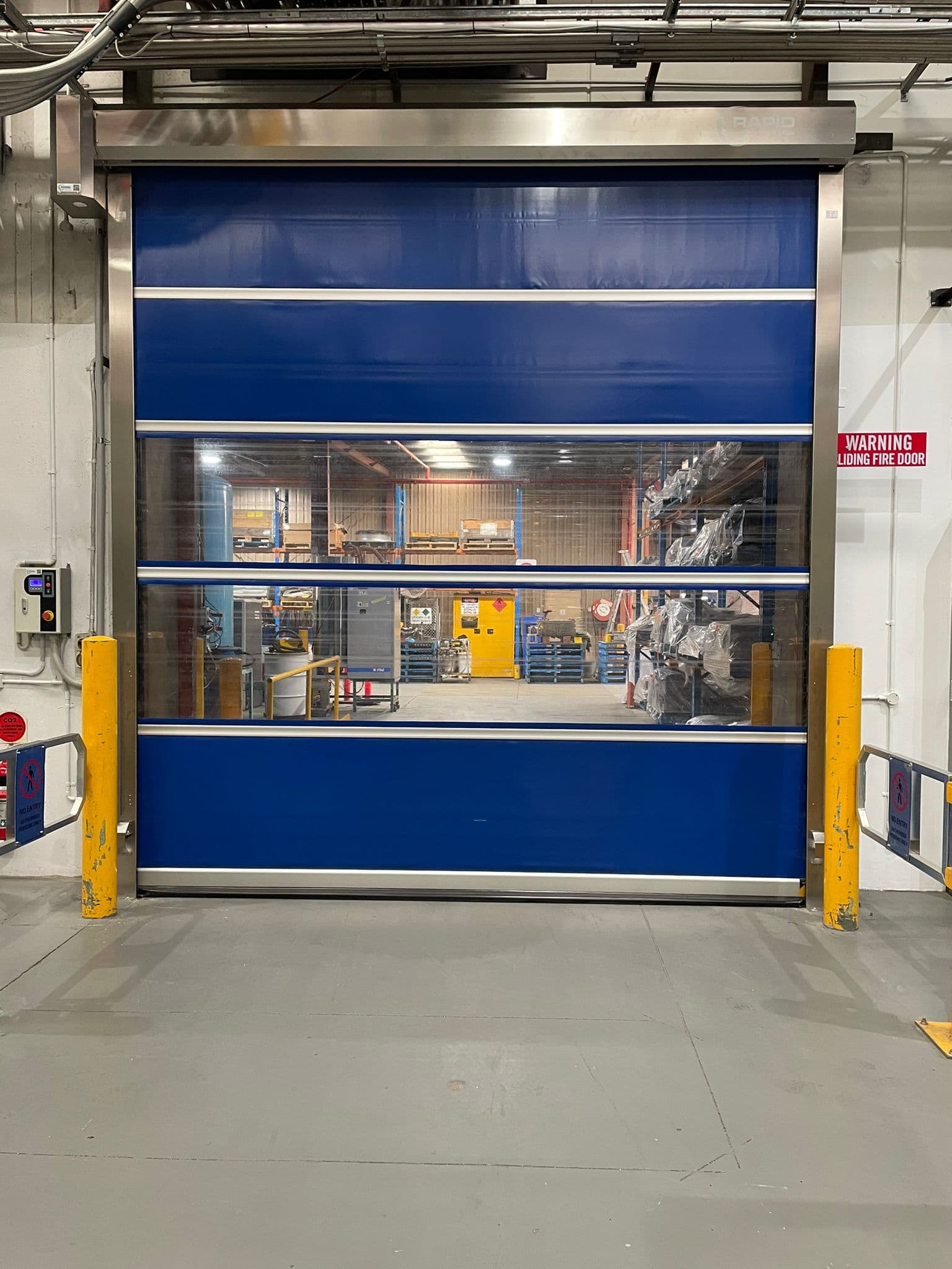 Blue rapid roll-up high-speed door with vision panels and bollards.