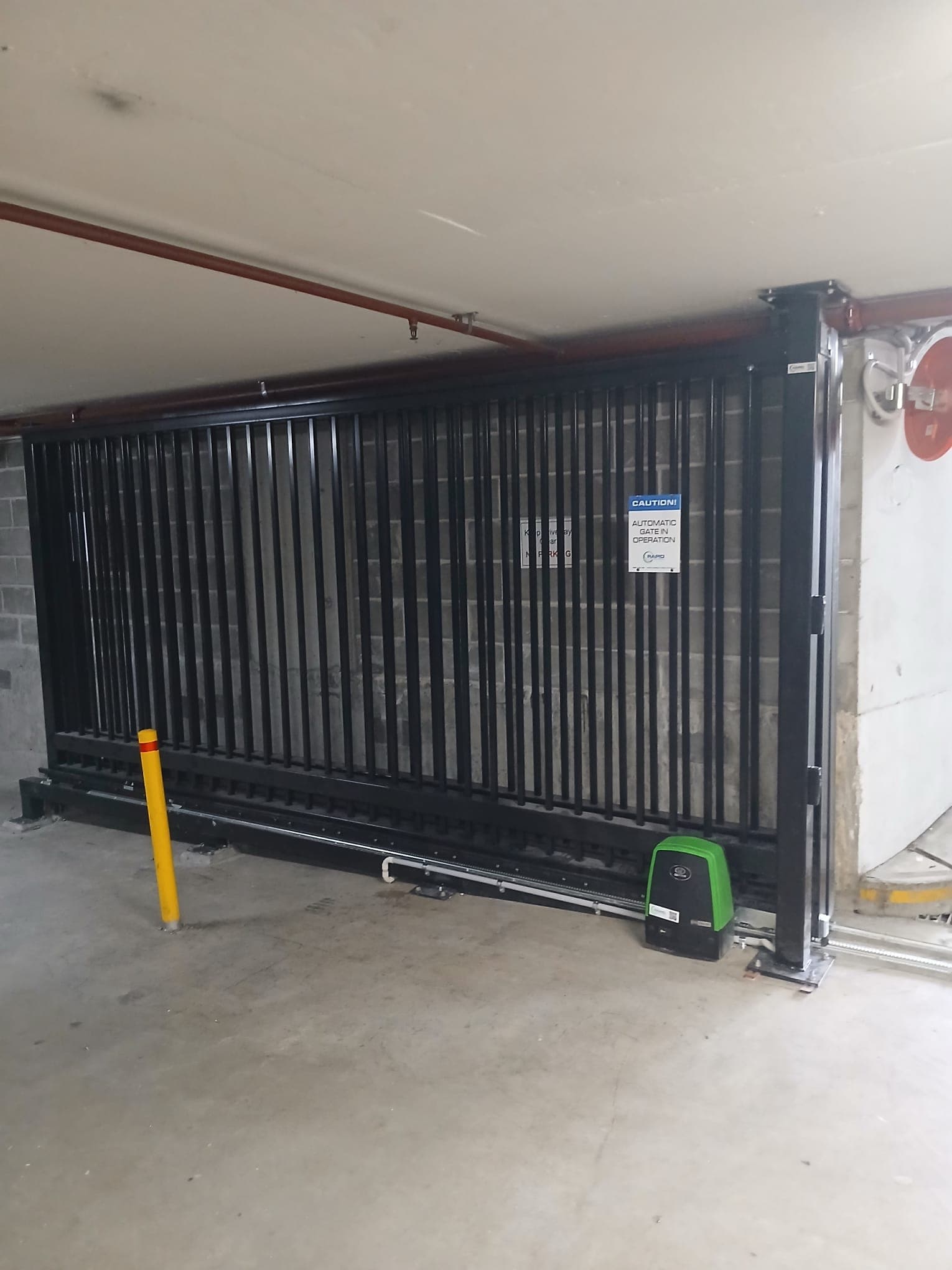 Black telescopic gate with motor unit, track, and safety bollard in an indoor car park.