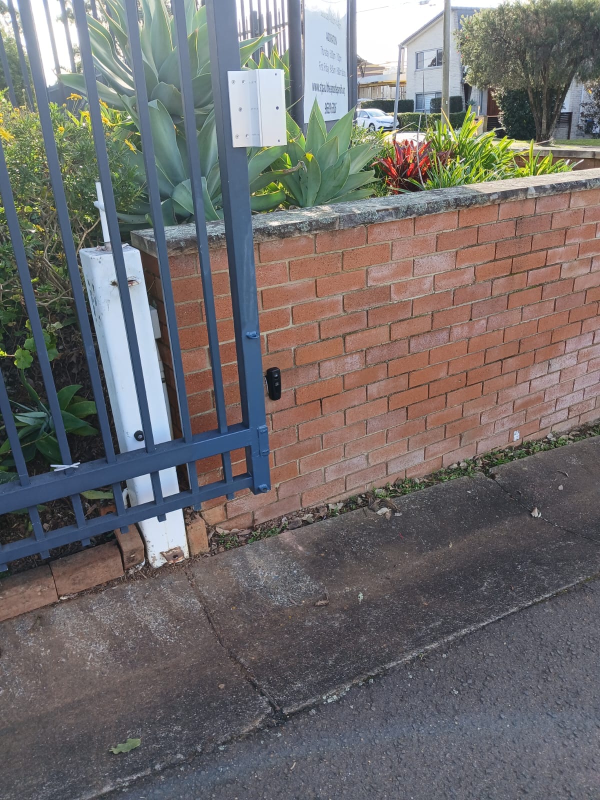 Dark grey custom vehicle access gate with control hardware beside a red brick boundary wall.